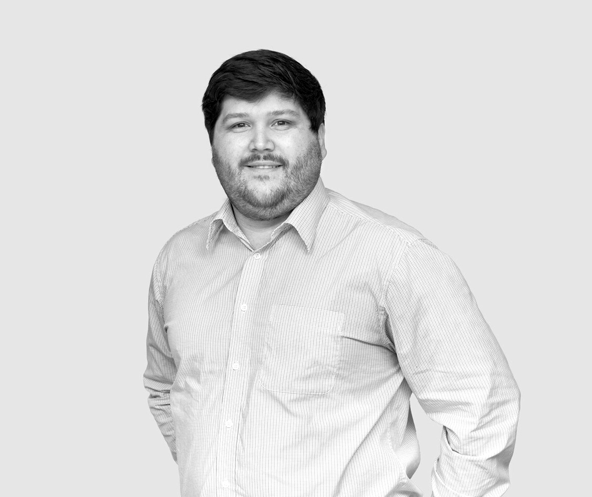 A black and white photo of a person with short dark hair and a beard, wearing a striped long-sleeved button-up shirt. They are standing against a plain background, facing the camera with a neutral expression.