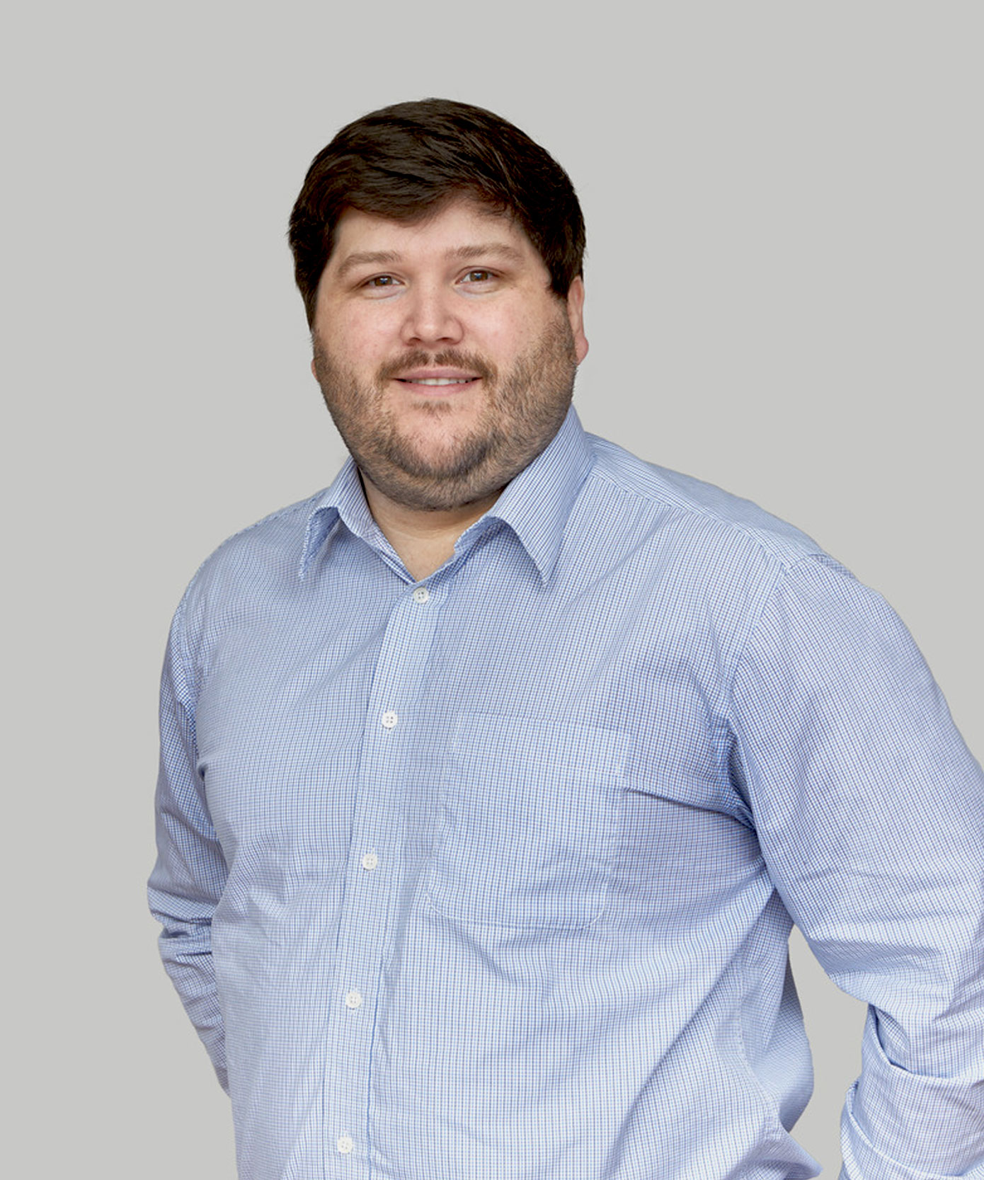 A person with short dark hair and a beard is wearing a light blue button-up shirt. They are standing in front of a plain gray background, looking at the camera with a neutral expression.